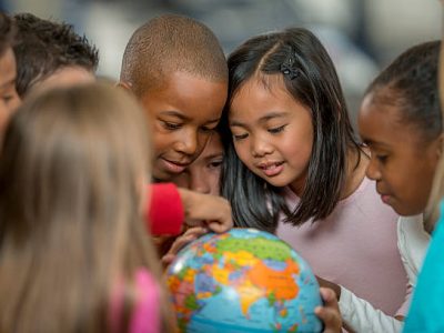 A multi-ethnic group of elementary age students are learning the places of the world by looking at a globe in geography and history class.