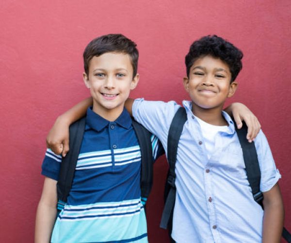 Best children friends standing with hand on shoulder against red background. Happy smiling classmates standing together on red wall after school. Portrait of multiethnic schoolboys enjoying friendship.